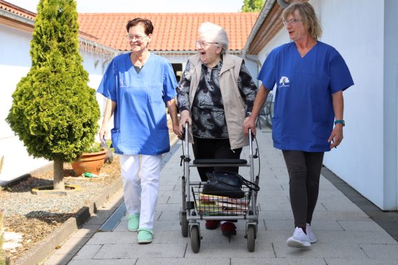 Fritz Reuter Pflegedienst GmbH Zwei Pflegende begleiten eine ältere Frau mit einem Rollator auf einem Gehweg.