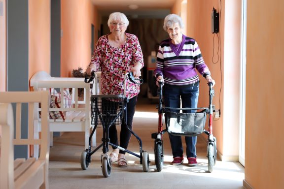 Fritz Reuter Pflegedienst GmbH Zwei ältere Frauen mit Gehhilfen gehen in einem Flur.