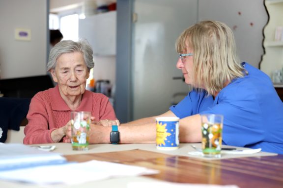 Fritz Reuter Pflegedienst GmbH Zwei Frauen sitzen am Tisch, eine ältere Frau hält die Hand der anderen. Gläser und Papiere liegen daneben.