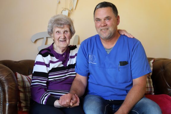 Fritz Reuter Pflegedienst GmbH Ältere Frau und ein Mann in blauer Arbeitskleidung sitzen auf einer Couch und lächeln.