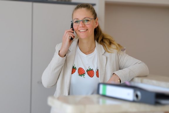 Fritz Reuter Pflegedienst GmbH Lächelnde Frau mit Telefon, trägt ein T-Shirt mit Erdbeer-Motiv, am Schreibtisch.