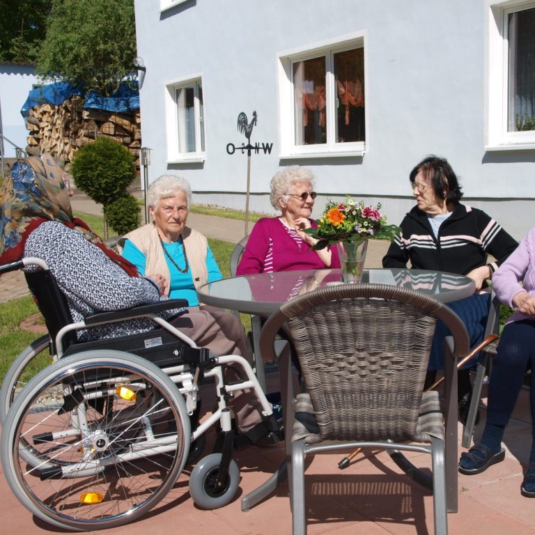 Fritz Reuter Pflegedienst Ältere Frauen sitzen am Tisch im Freien, einige im Rollstuhl, bei schönem Wetter.