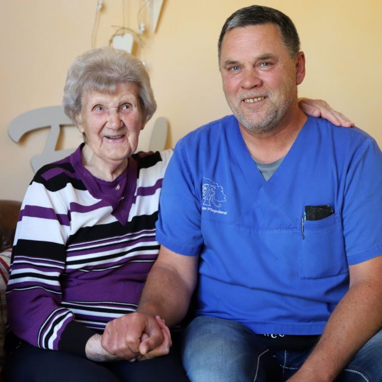 Fritz Reuter Pflegedienst Ältere Frau und Mitarbeiter in blauer Arbeitskleidung sitzen lächelnd zusammen.