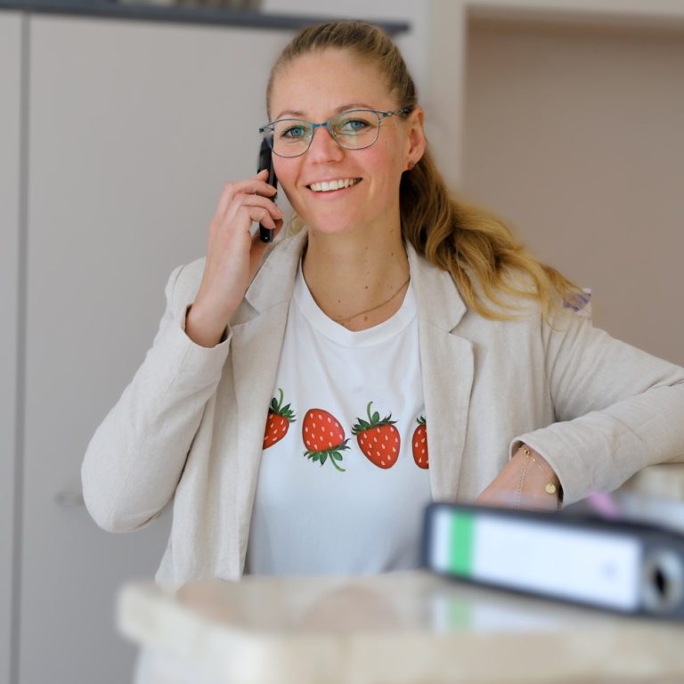 Nancy Schwindeler – Prokuristin Frau mit blonden Haaren und Brille, am Telefon, trägt T-Shirt mit Erdbeeren.
